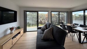 Ein modernes Wohnzimmer mit einer Couch, einem Esstisch und Stühlen. Ein Fernseher ist an der Wand befestigt und ein Fenster bietet einen Blick ins Freie.