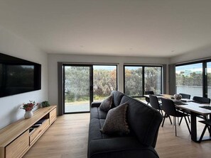 A modern living room with a sofa, dining table, and chairs. There is a TV mounted on the wall and a window providing a view of the outdoors. - Wave Front - Freycinet Holiday Houses (COLES BAY)