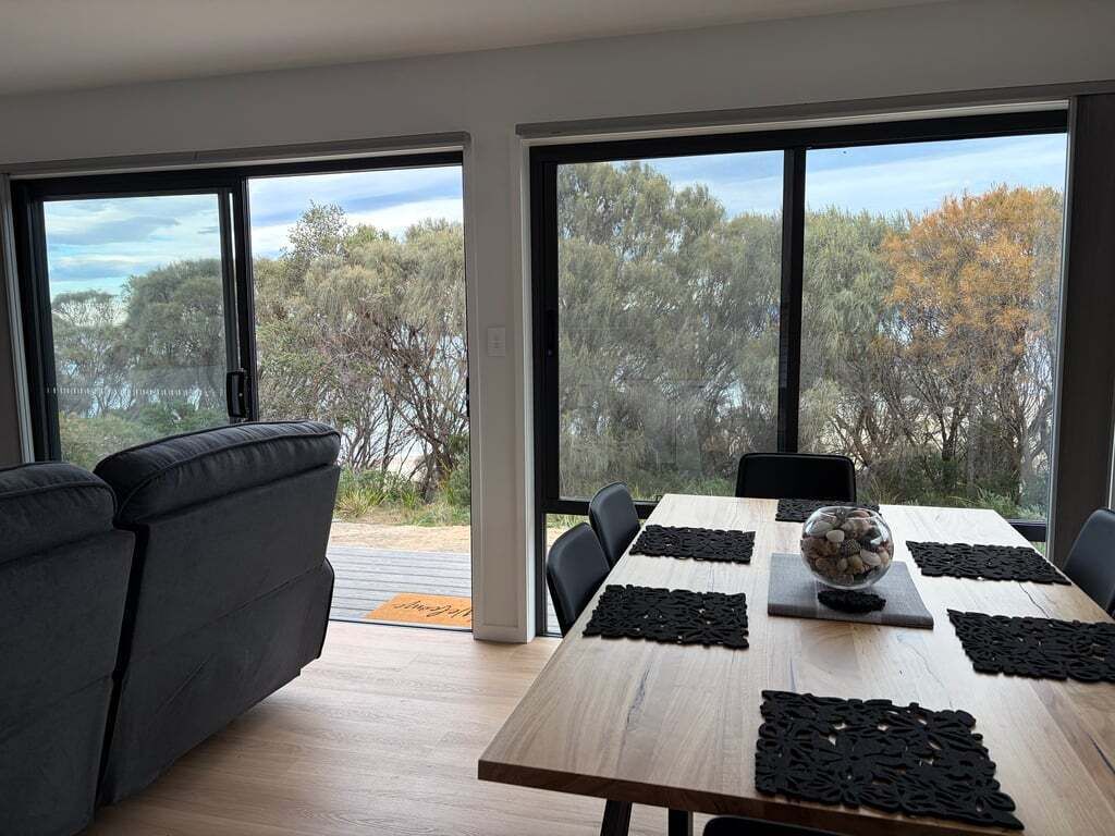 Een moderne eetruimte met een houten tafel, zwarte placemats en een schaal met stenen. Grote ramen bieden uitzicht op bomen en een wateroppervlak.