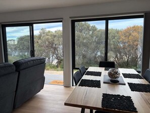 A modern dining area with a wooden table, black placemats, and a bowl of stones. Large windows offer a view of trees and a body of water. - Wave Front - Freycinet Holiday Houses (COLES BAY)