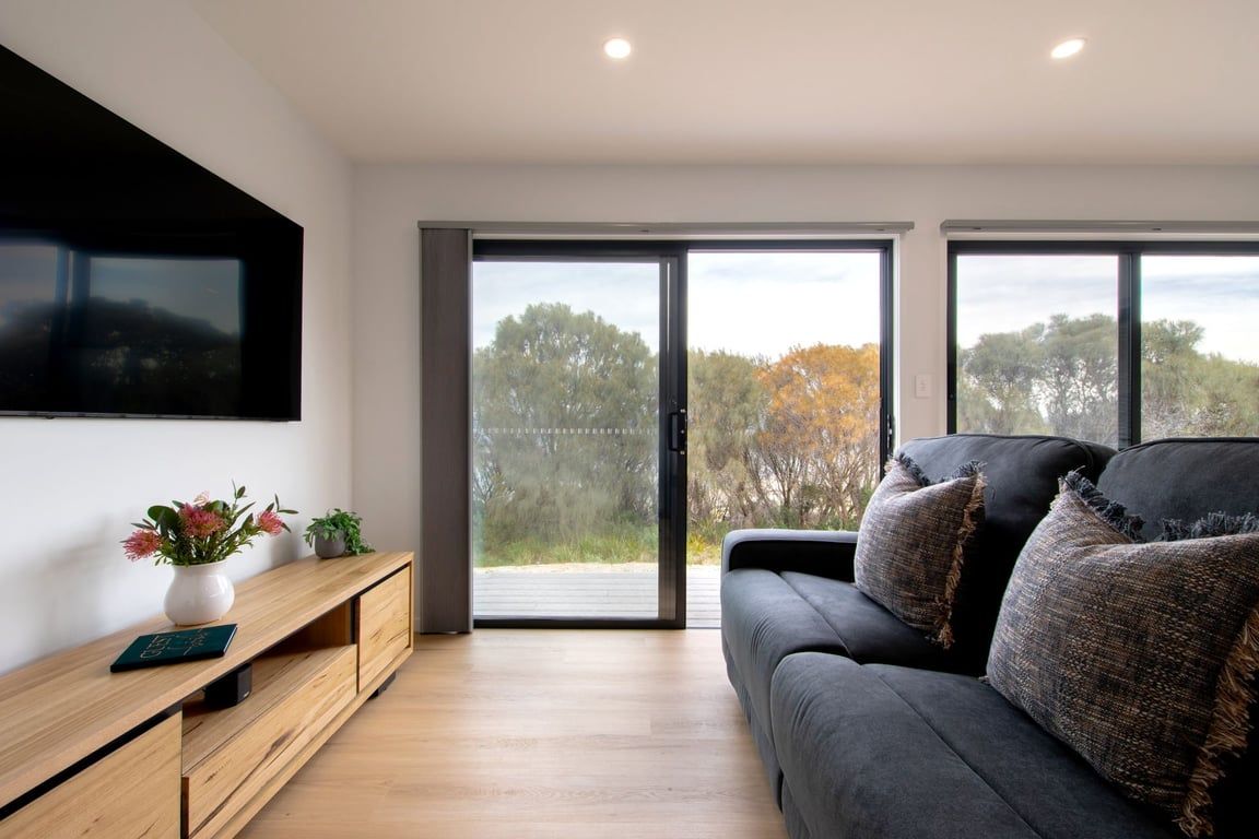 A modern living room with a flat-screen TV mounted on the wall, a wooden console table with a vase and plants, a dark grey sofa with textured cushions, and large sliding glass doors leading to an outdoor area.