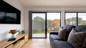 A modern living room with a flat-screen TV mounted on the wall, a wooden console table with a vase and plants, a dark grey sofa with textured cushions, and large sliding glass doors leading to an outdoor area.