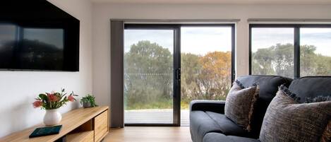 A modern living room with a flat-screen TV mounted on the wall, a wooden console table with a vase and plants, a dark grey sofa with textured cushions, and large sliding glass doors leading to an outdoor area.