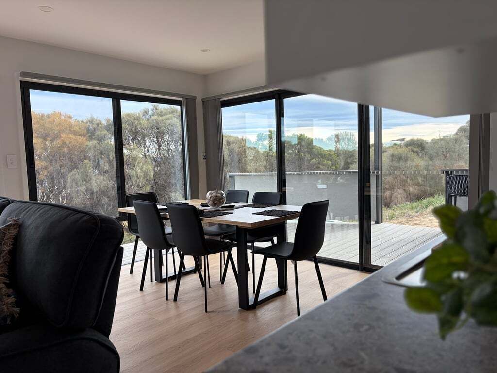 Et moderne spiseområde med et bord og stole, store vinduer med udsigt til træer og et vandareal, samt en balkon med grill.