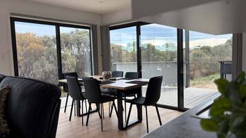 Et moderne spiseområde med et bord og stole, store vinduer med udsigt til træer og et vandareal, samt en balkon med grill.