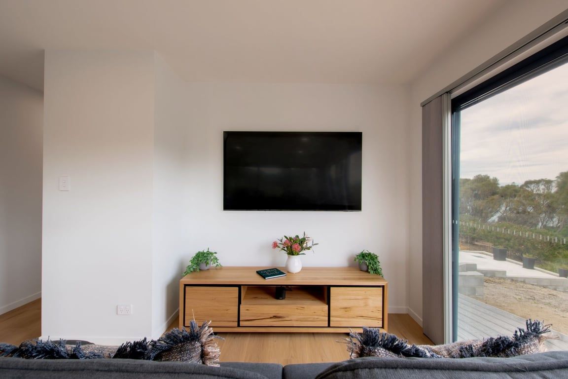 A modern living room with a flat-screen TV mounted on the wall, a wooden entertainment unit with a vase of flowers, and a large window offering a view of the outdoors.