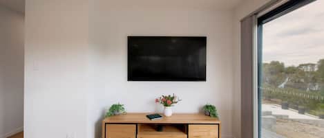A modern living room with a flat-screen TV mounted on the wall, a wooden entertainment unit with a vase of flowers, and a large window offering a view of the outdoors.