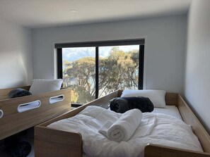 A bedroom with a large window, a bed with white bedding, and a wooden headboard. - Wave Front - Freycinet Holiday Houses (COLES BAY)