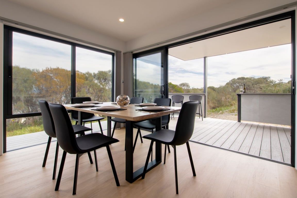 A modern dining area with a large table and chairs, large windows, and a view of the outdoors.