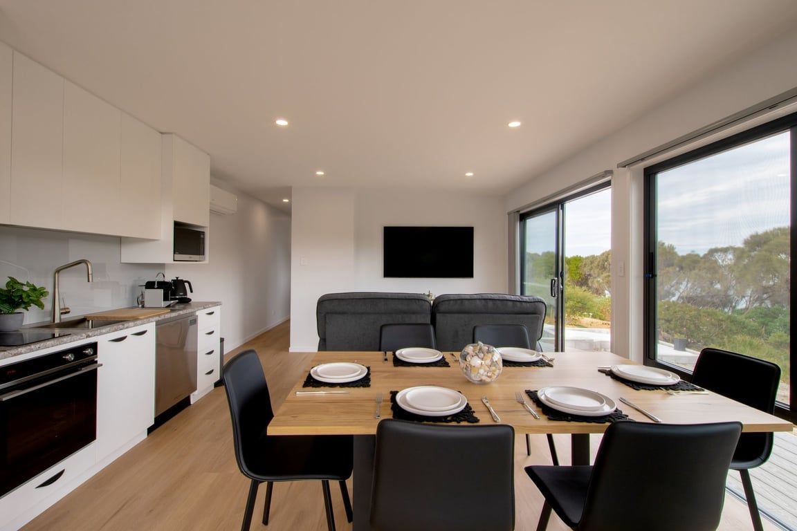 Een moderne keuken met witte kasten, een inbouwoven en een eethoek met een houten tafel voor vier personen.