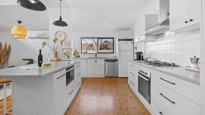 A modern kitchen with white cabinetry, a central island, a stove, and a window with a view of trees. - House on Drummond - A large home close to the heart of central Ballarat (Ballarat Central)