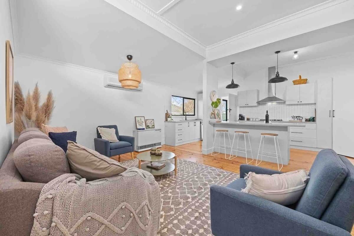 Una sala de estar moderna con un sofá, un sillón y una mesita de centro. Se observa una zona de cocina con barra y taburetes.