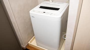 A compact, white, wall-mounted washing machine in a corner of a room with a wooden floor and beige walls.