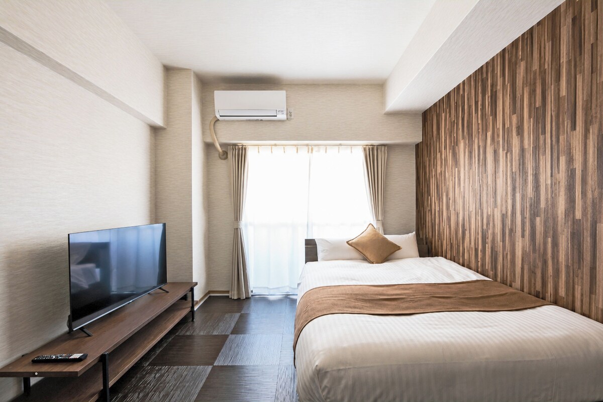 A hotel room with a bed, a TV on a stand, a window with curtains, and a wall-mounted air conditioning unit.