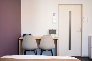 Habitación de hotel con cama, escritorio con computadora, dos sillas, una puerta y un termostato en la pared.