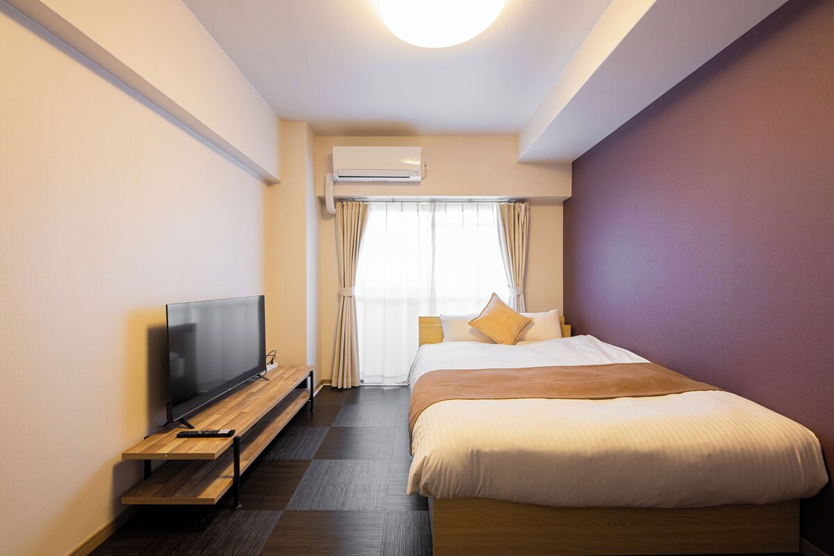 A hotel room with a bed, a TV on a wooden stand, a window with curtains, and an air conditioning unit.