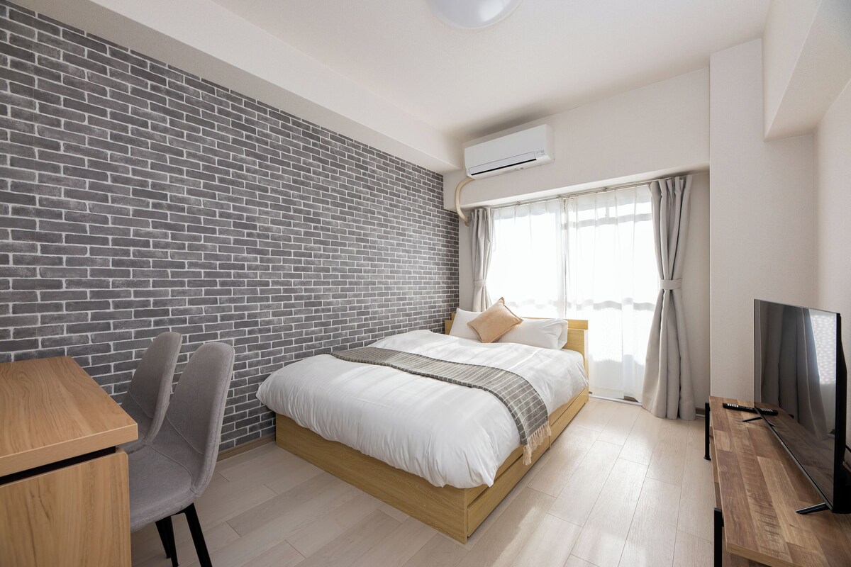 A modern hotel room with a brick wall, a bed with white bedding, a desk with a chair, a TV, and a window with curtains.