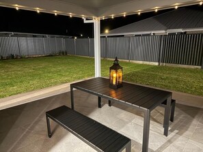 An outdoor patio with a table and bench, a lantern, and a fenced yard at night.