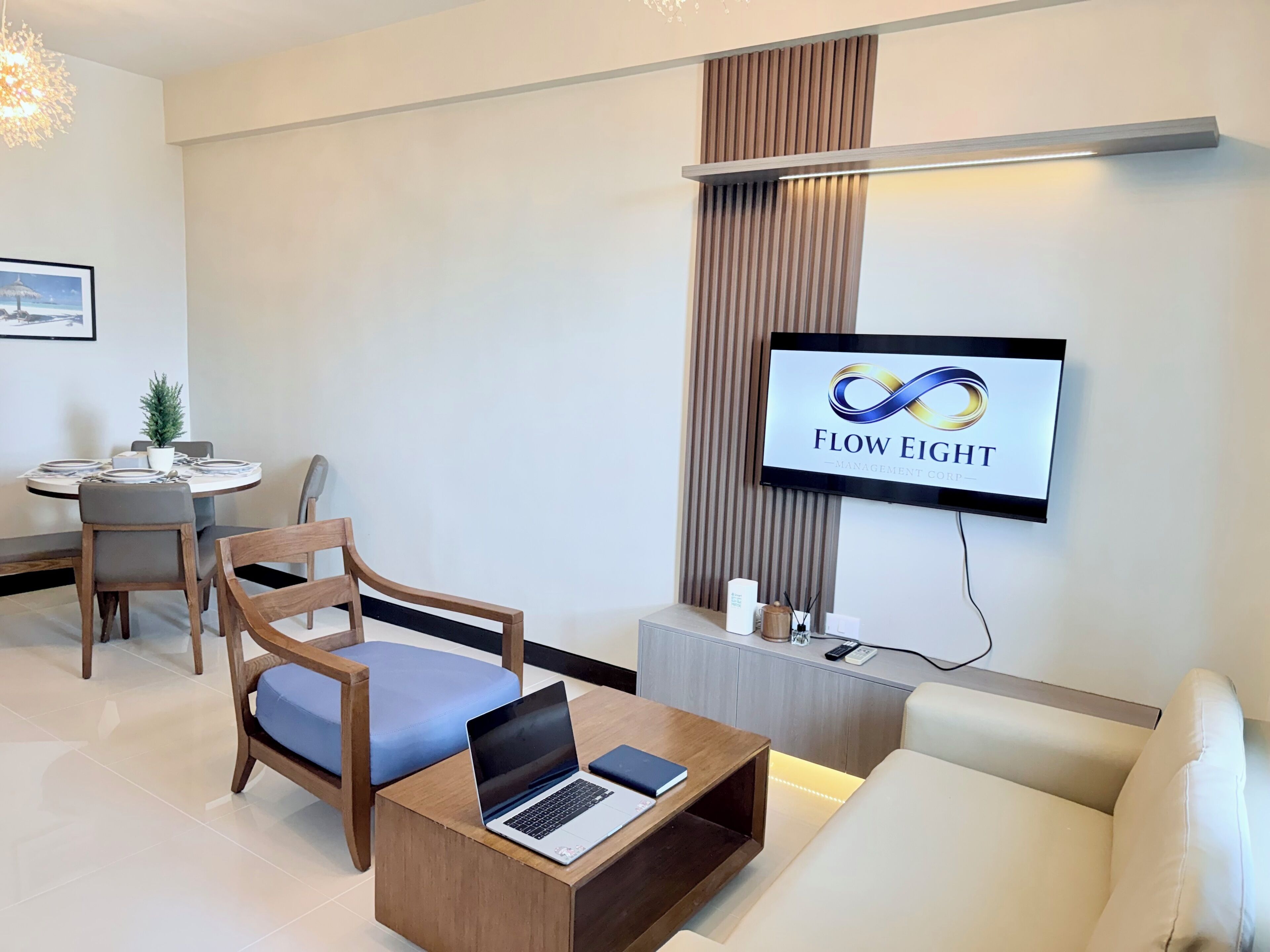 A modern living room with a sofa, a coffee table with a laptop, a TV mounted on the wall, and a dining area with a table and chairs.