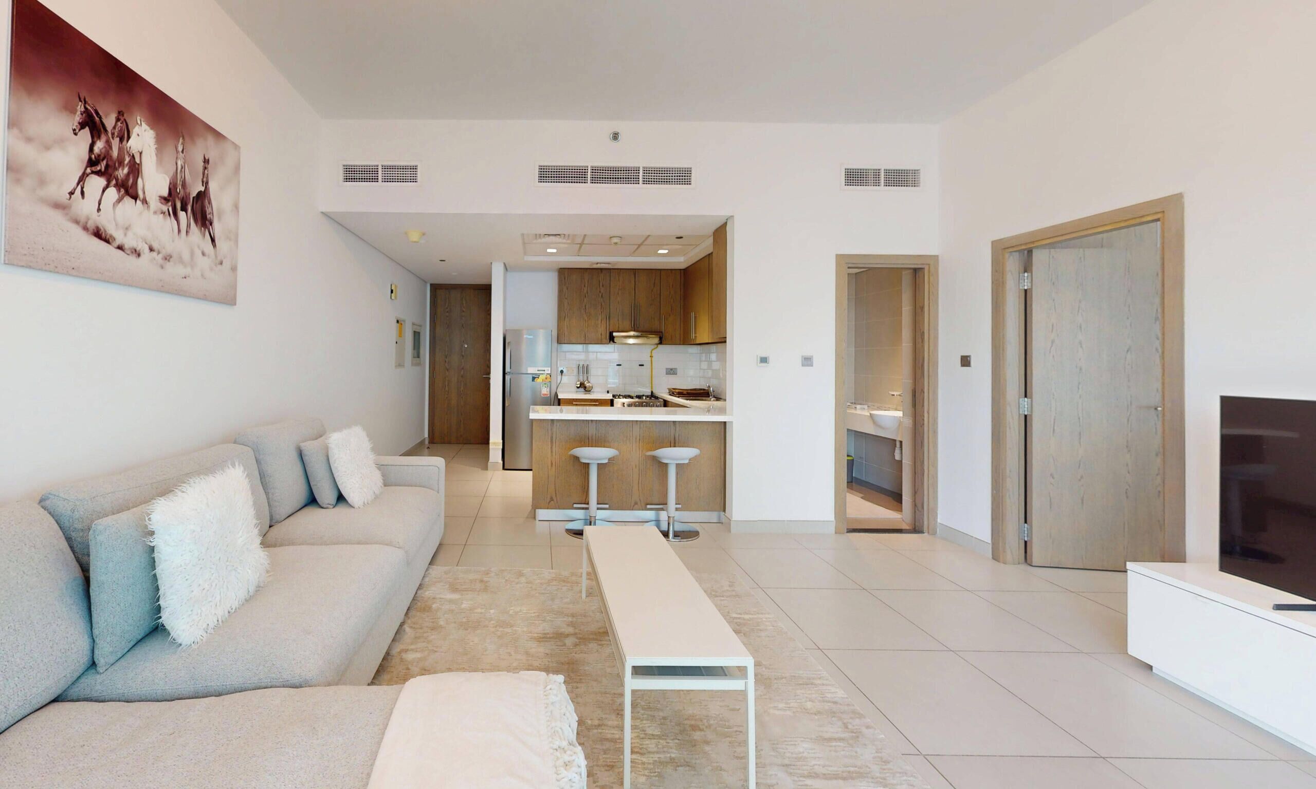 A modern living room with a sofa, a coffee table, and a TV. There is a kitchen in the background with wooden cabinets and a dining area.