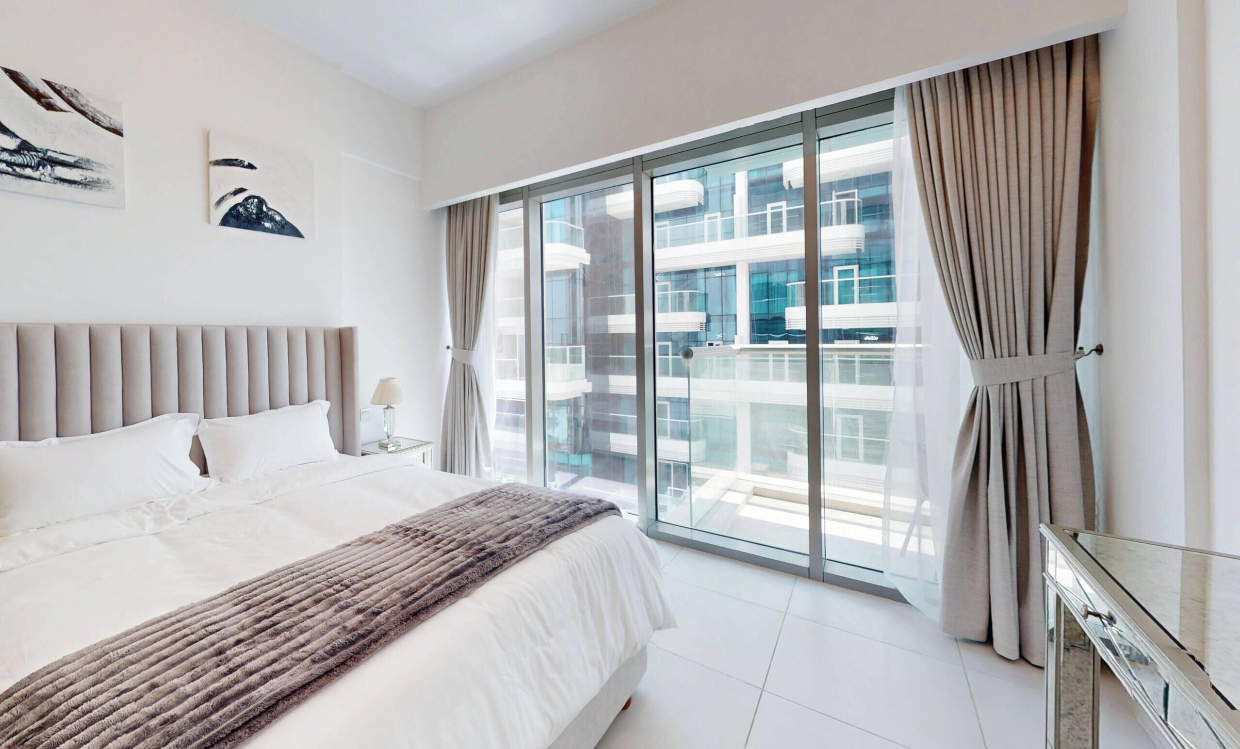 Quarto de hotel moderno com uma cama grande, vista de um edifício através de janelas do chão ao teto e uma varanda com cortinas.