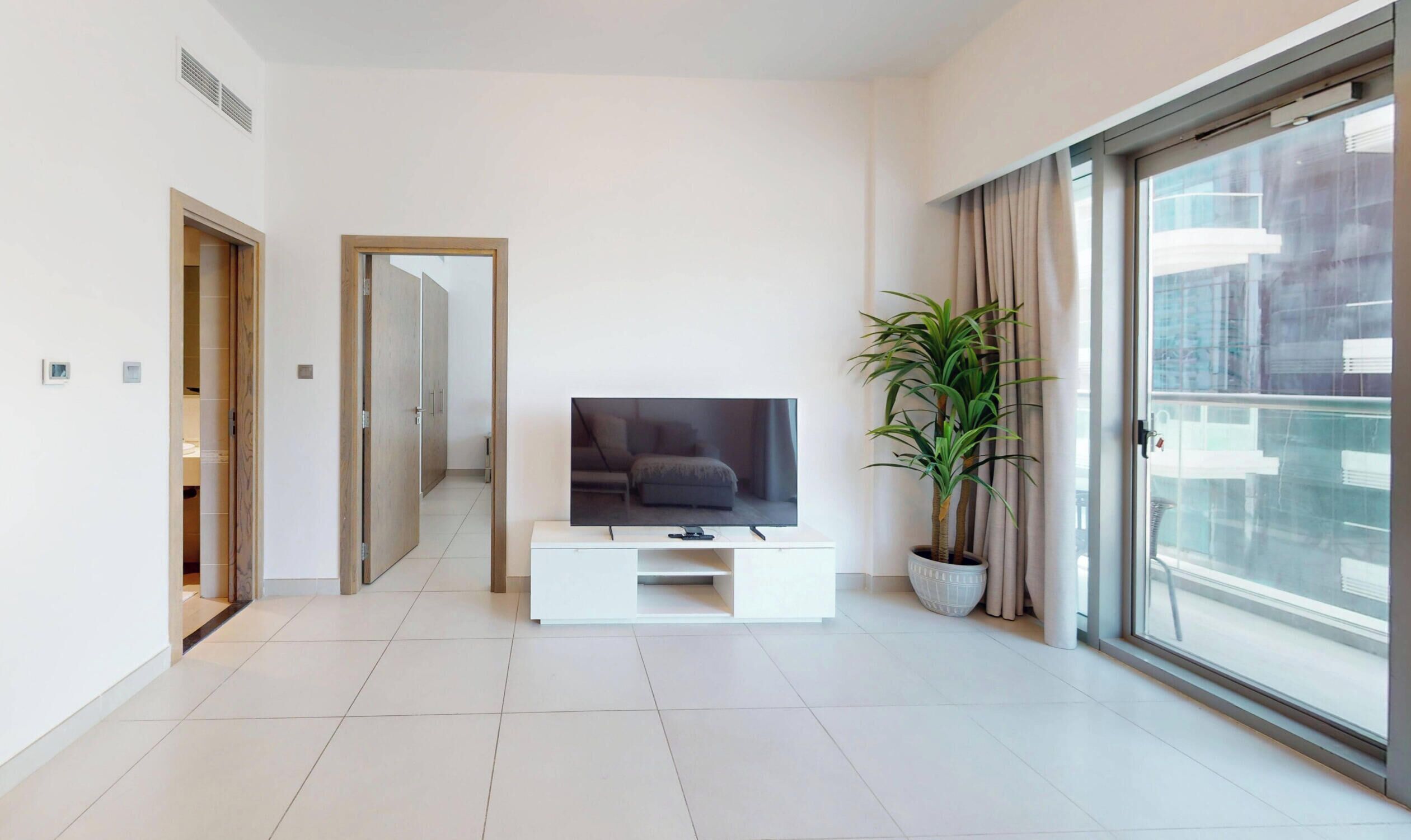 A modern living room with a flat-screen TV on a white console, a potted plant, and a large window with a view of the city.