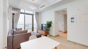 A modern living room with a sofa, a dining table, and a TV. There is a large window with a view of the city. - Chic 1BR with Canal View (Dubai)