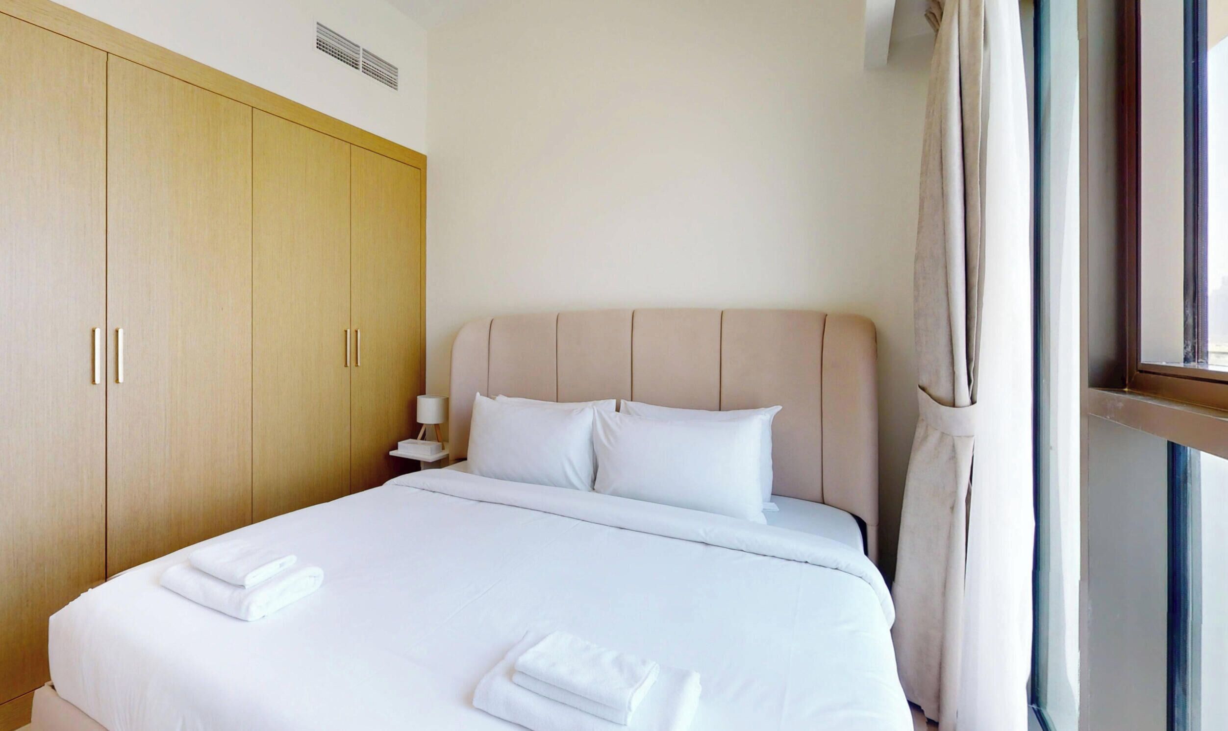 Una habitación de hotel con una cama grande, un armario de madera, una mesita de noche con lámpara y una ventana con cortinas.