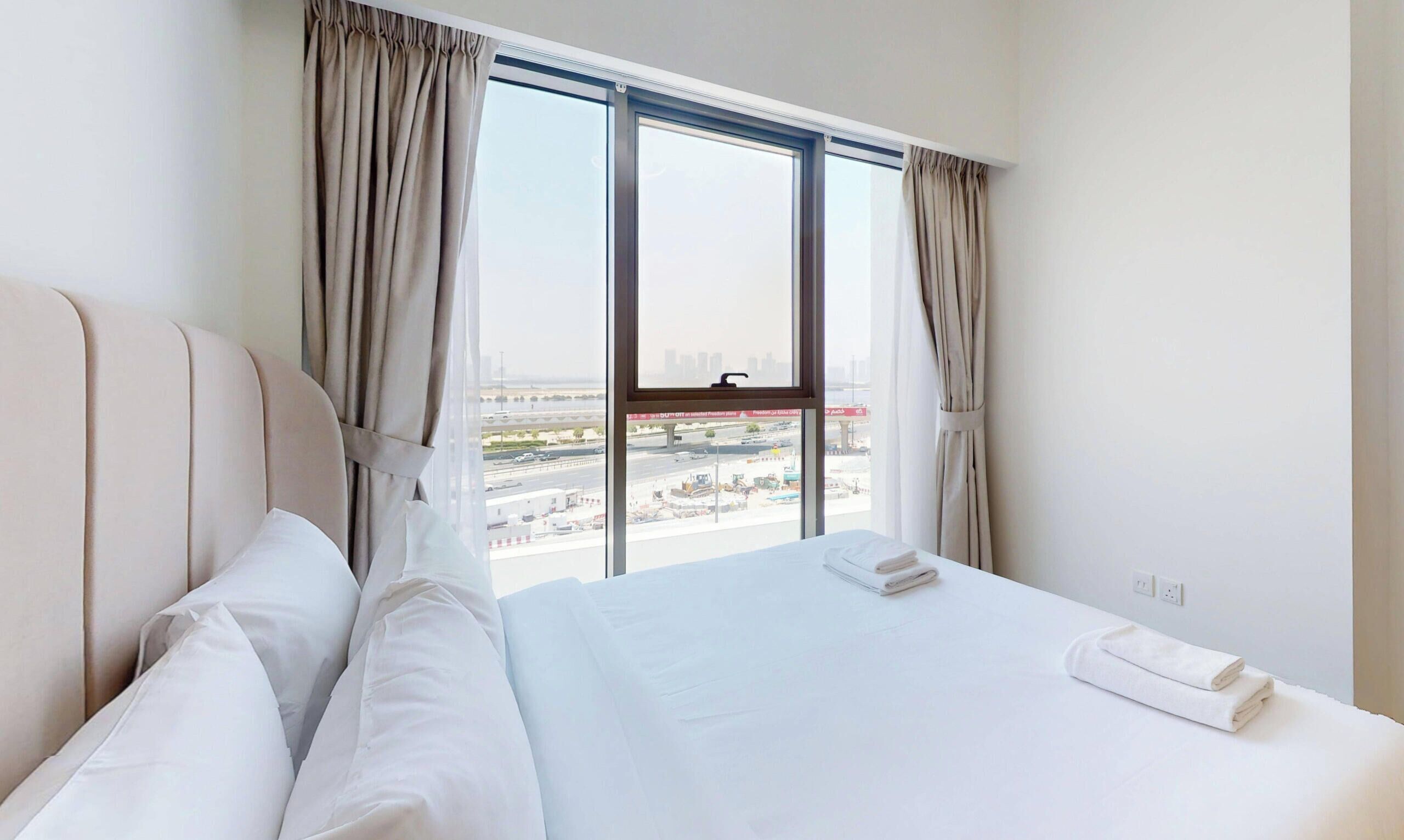 A hotel room with a large bed, two pillows, a neatly made bed with a folded towel, and a view of a cityscape through a large window with curtains.