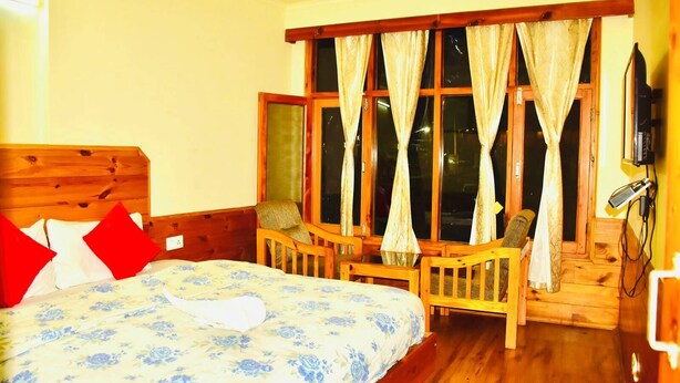 A room with a bed, a wooden bench, a chair, a window with curtains, and a television.