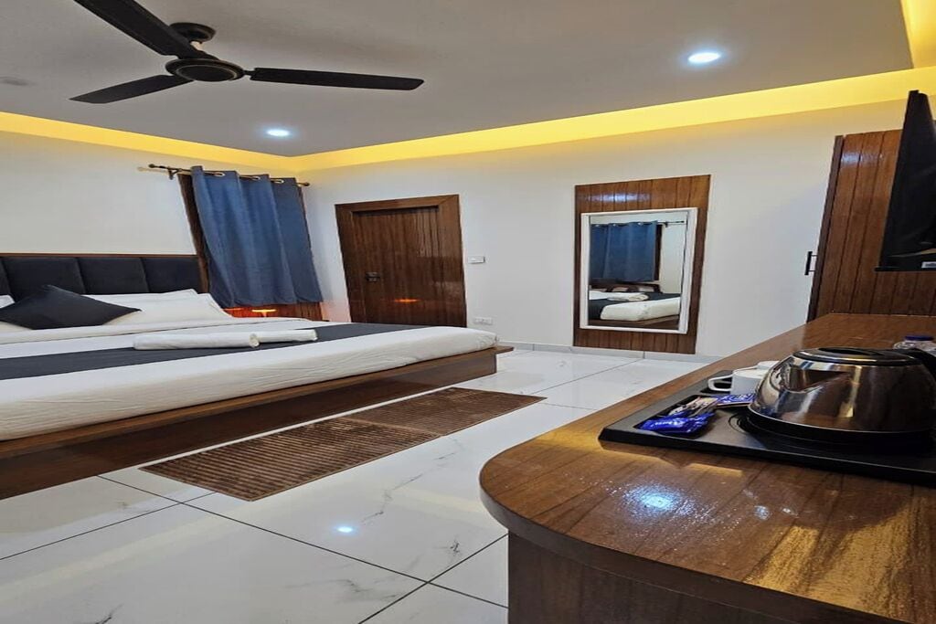 A hotel room with a bed, a desk with a kettle, a ceiling fan, and a mirror.