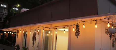 A building with a covered outdoor area, potted plants, and hanging light bulbs.