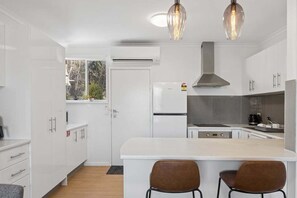 A modern kitchen with white cabinetry, a central island with two chairs, and pendant lighting. - Snowy Nook (Jindabyne)