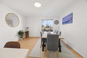 A dining area with a long table, chairs, a mirror, a clock, and a window with a view. - Snowy Nook (Jindabyne)