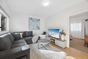 A modern living room with a grey sectional sofa, a flat-screen TV, a small round coffee table, and a white shag rug. - Snowy Nook (Jindabyne)