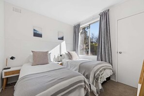 A bedroom with two beds, a nightstand, a lamp, and a window with curtains. - Snowy Nook (Jindabyne)