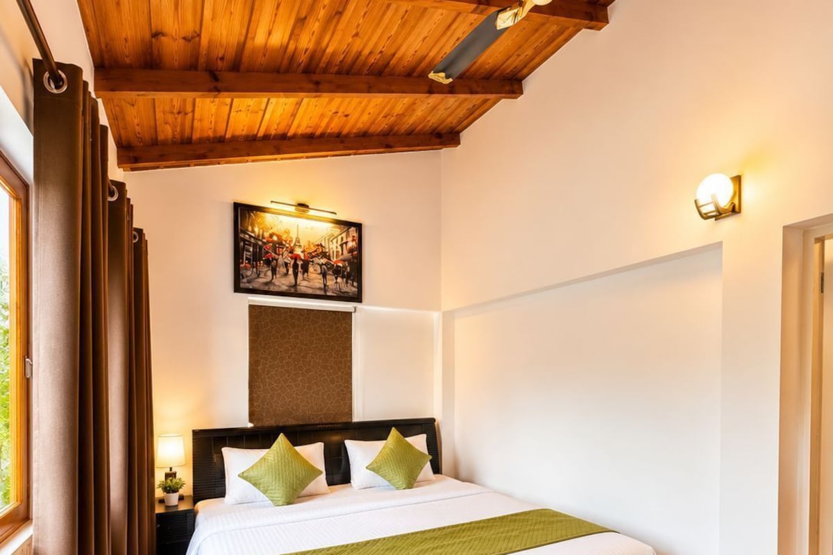 A bedroom with a wooden ceiling, a bed with a green throw, a nightstand with a lamp, and a wall-mounted picture.