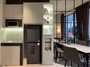 A compact kitchen with a built-in microwave, a refrigerator, and a dining area with a table and chairs. - LaBriza By SanLiving - 2BR - PIK2 (Kosambi)