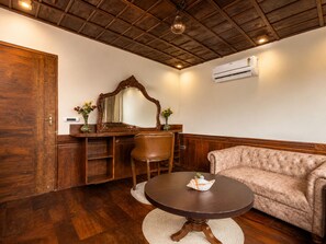 A room with a wooden ceiling, a round table with a decorative shell, a cushioned sofa, a wooden desk with a mirror, and an air conditioning unit.