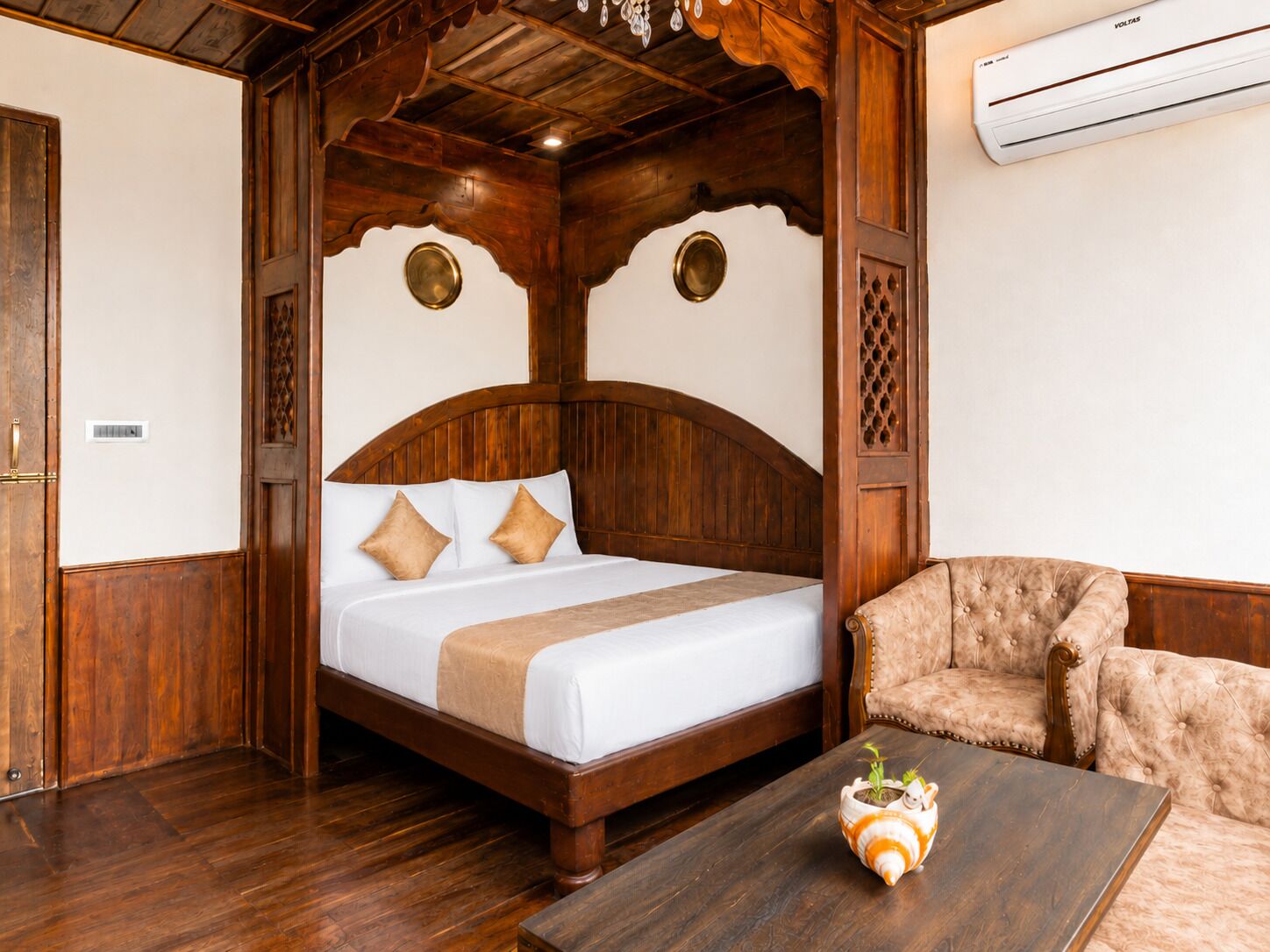 Una cama con estructura de madera, colchón blanco y almohadas color beige, un sillón capitonado y una mesita con una planta.