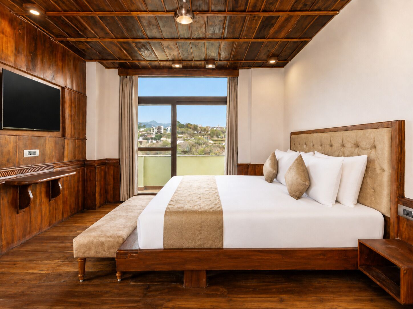 Habitación de hotel con techo de madera, una cama grande con ropa de cama blanca y beige, un televisor de pantalla plana montado en la pared y vistas al exterior a través de un ventanal.