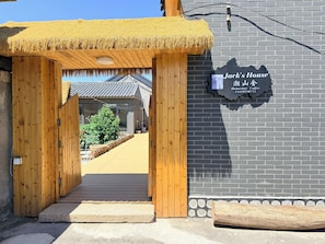 A wooden entrance with a thatched roof leading to a building with a brick facade and a sign reading 'Jack's House Homestay Coffee.'