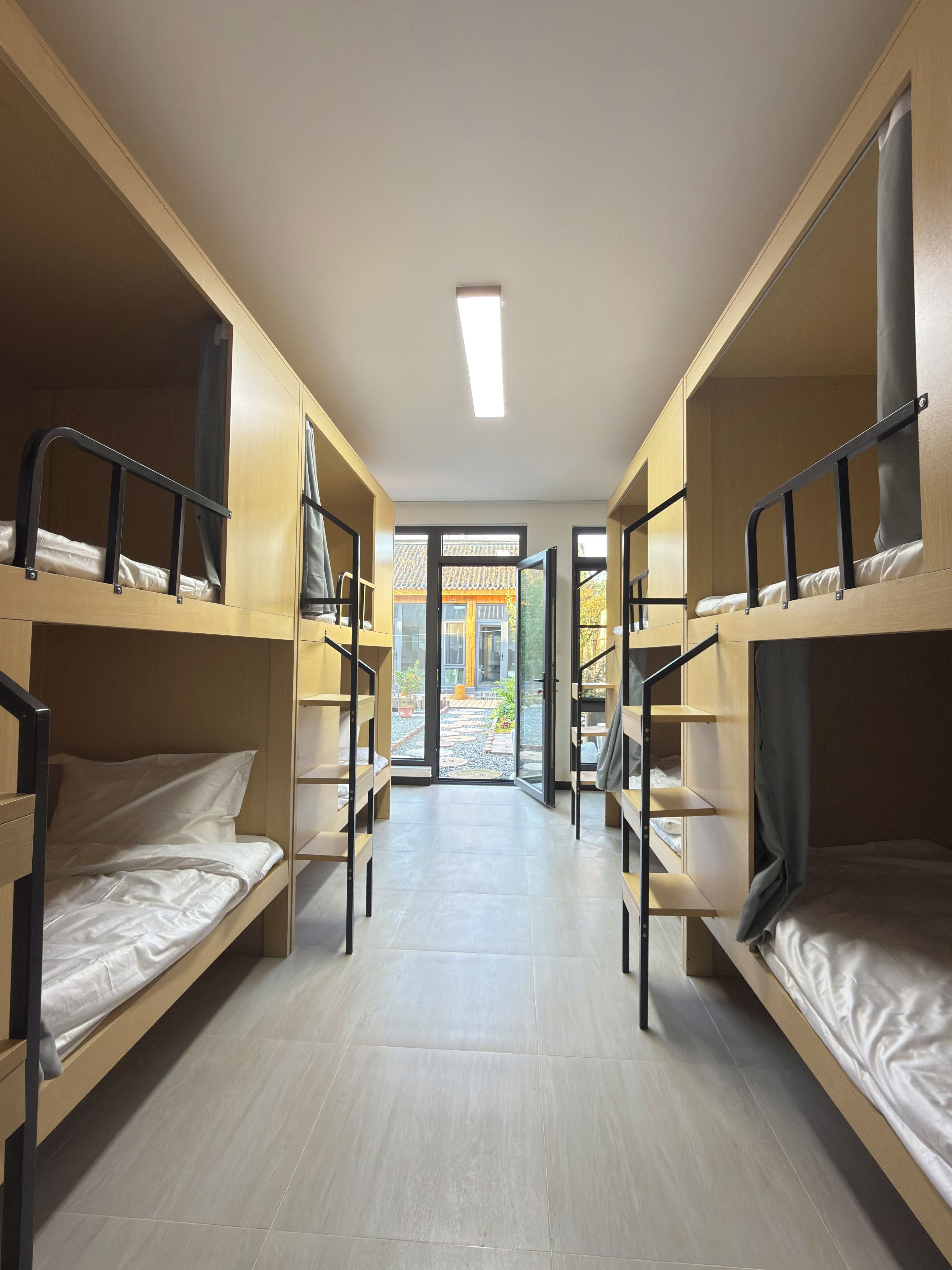 A corridor with bunk beds, a tiled floor, and a view of an outdoor area with a building and a garden.