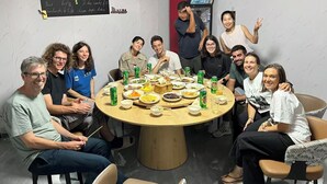 A group of people around a round table with food and drinks, in a room with a white wall and a mounted TV.