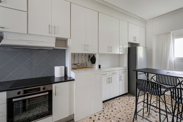 A modern kitchen with white cabinets, a black countertop, a built-in oven, and a stainless steel refrigerator. - Thriasion 10 Atelier (Athens)