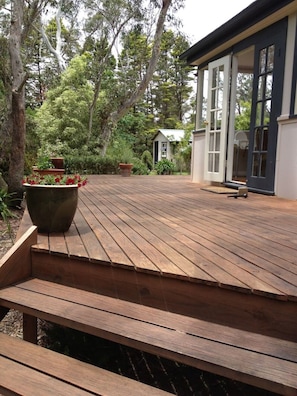 Eine Holzterrasse mit einer Bank, umgeben von üppigem Grün und einem kleinen Gebäude mit großen Fenstern.