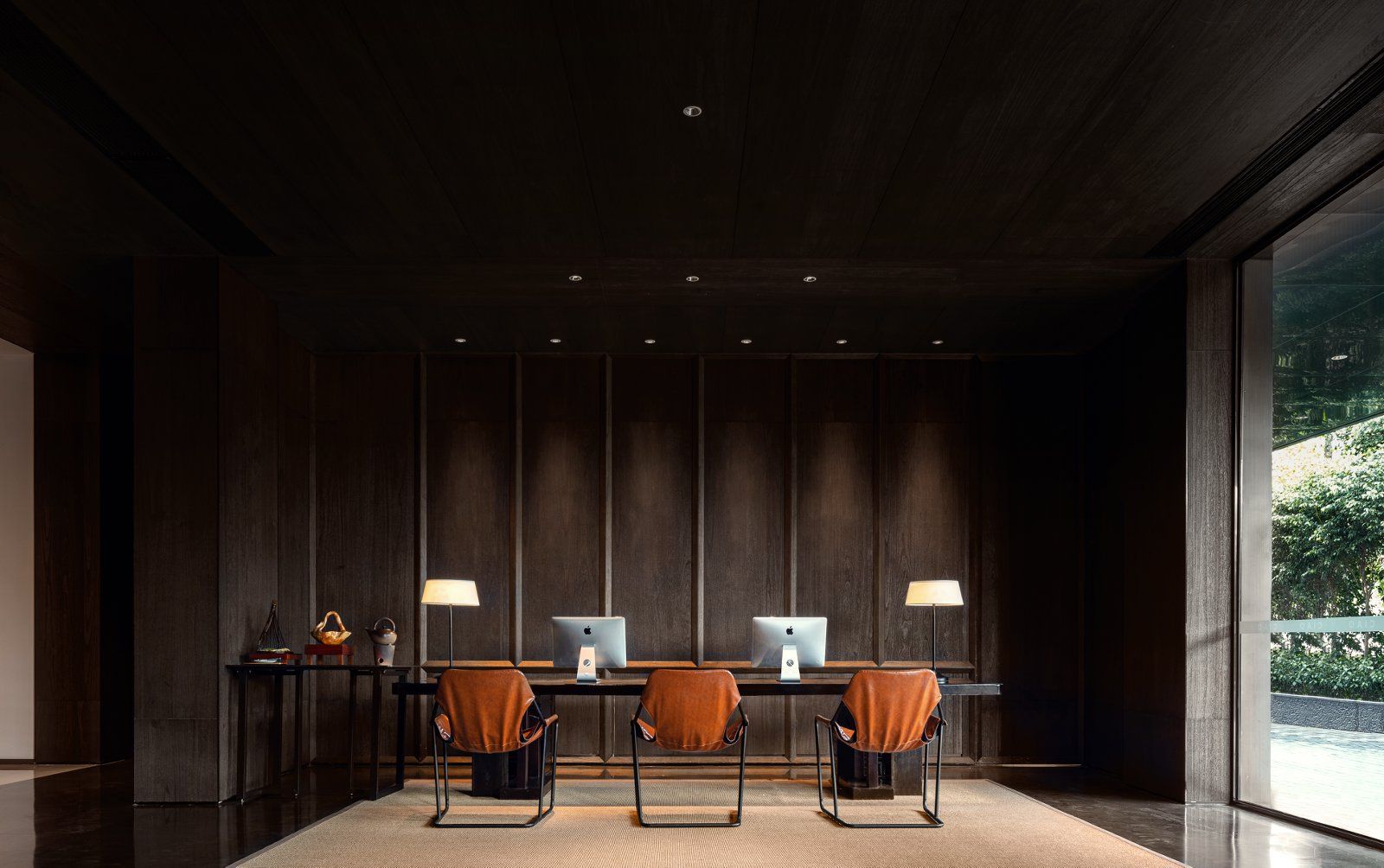 Una sala de conferencias moderna con un panel de pared de madera oscura, una larga mesa con dos Apple iMac, dos sillones de cuero y dos lámparas de pie.