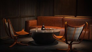 Una habitación oscura con un sofá de cuero, dos sillas de madera y una mesa redonda con una botella y vasos.