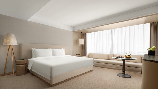 Habitación de hotel con una cama grande, un sofá, una mesita con una cesta, una lámpara y una ventana con cortinas.