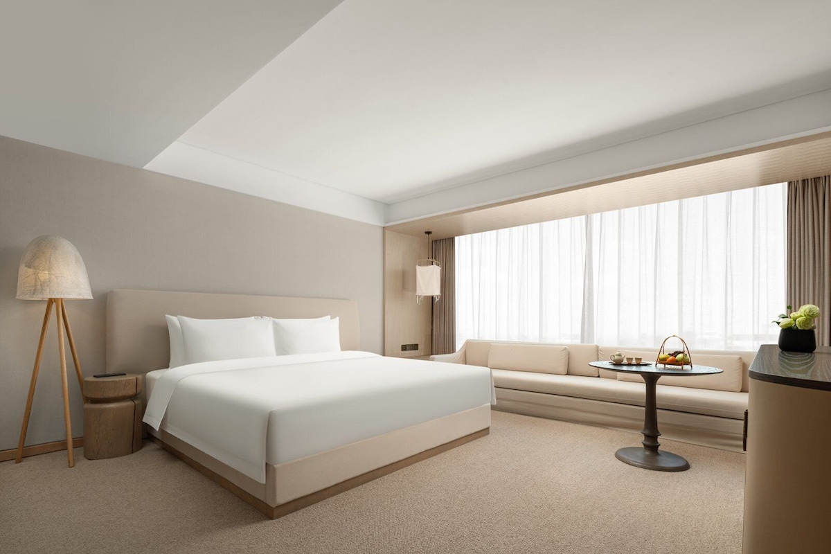 Habitació d'hotel amb un llit gran, un sofà, una taula petita amb una cistella, una làmpada i una finestra amb cortines.